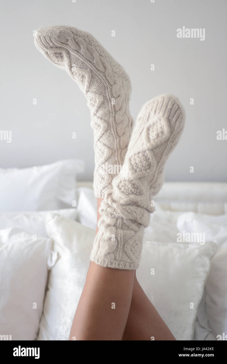 Par de piernas femeninas vistiendo calcetines crema acogedor tumbado en la cama con los pies en el aire con almohadas blancas detrás Foto de stock