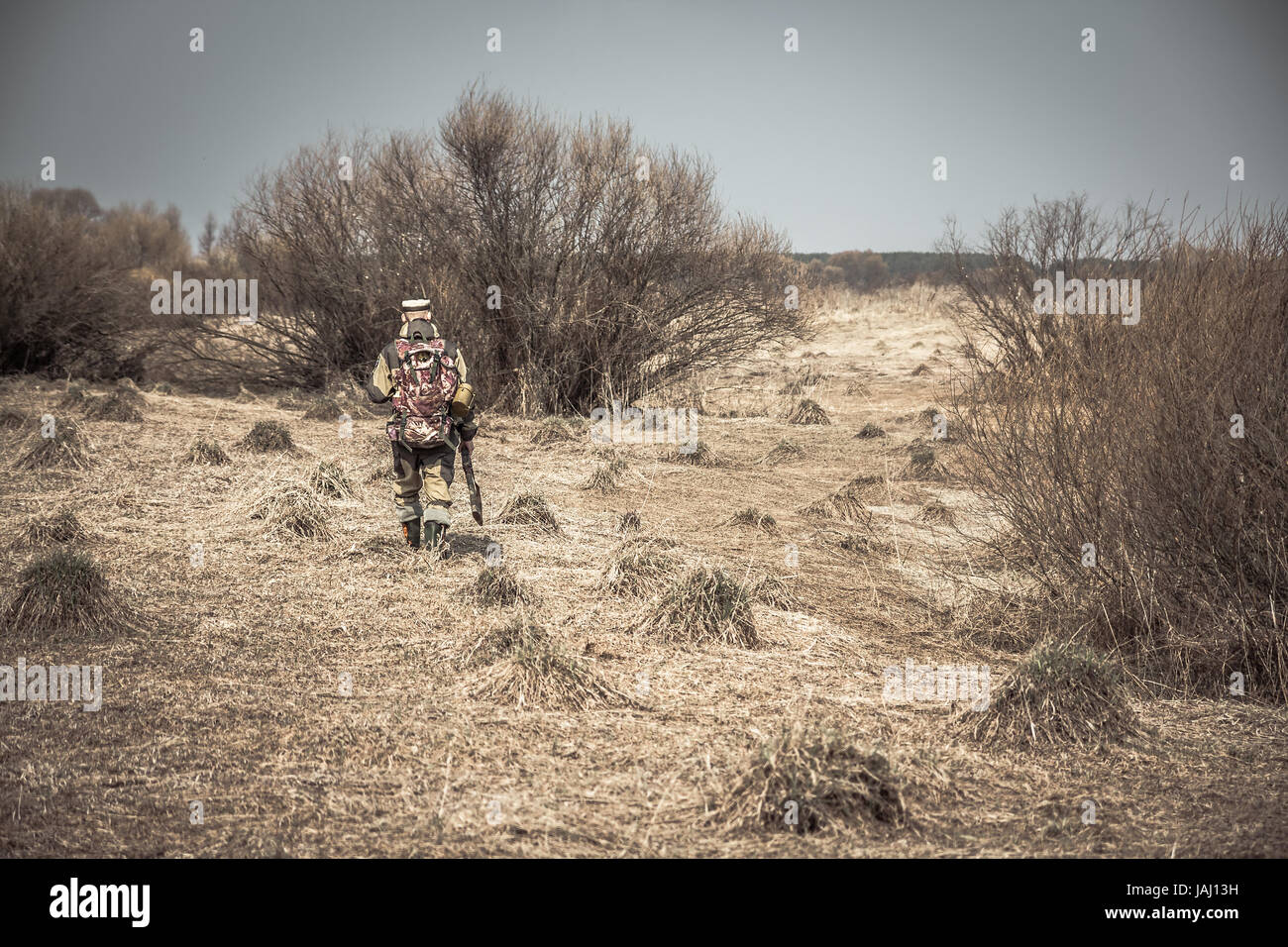 Hombre cazador de camuflaje con pistola atravesando la zona rural con pasto seco y arbustos durante la caza Foto de stock