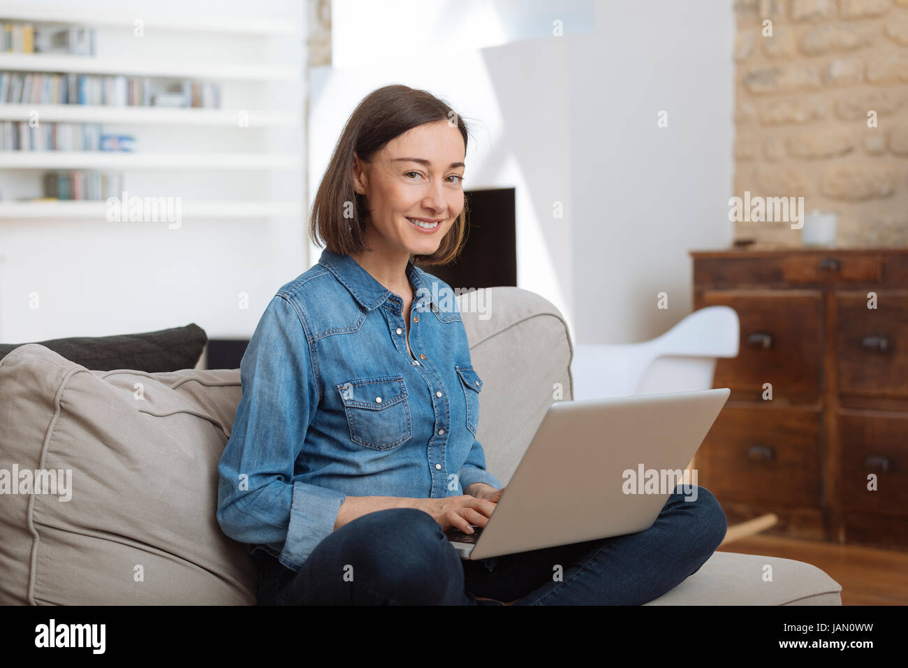 Atractiva mujer madura, usando un ordenador portátil mientras se relaja en un sofá en casa Foto de stock