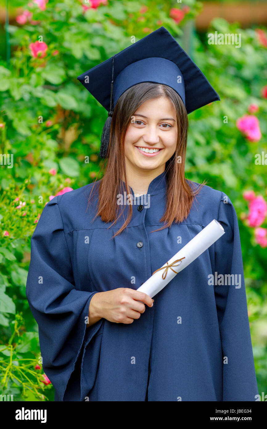Mujer sonriente sosteniendo el diploma y vistiendo toga y birrete afuera mirando a la cámara. Concepto de graduación. Foto de stock