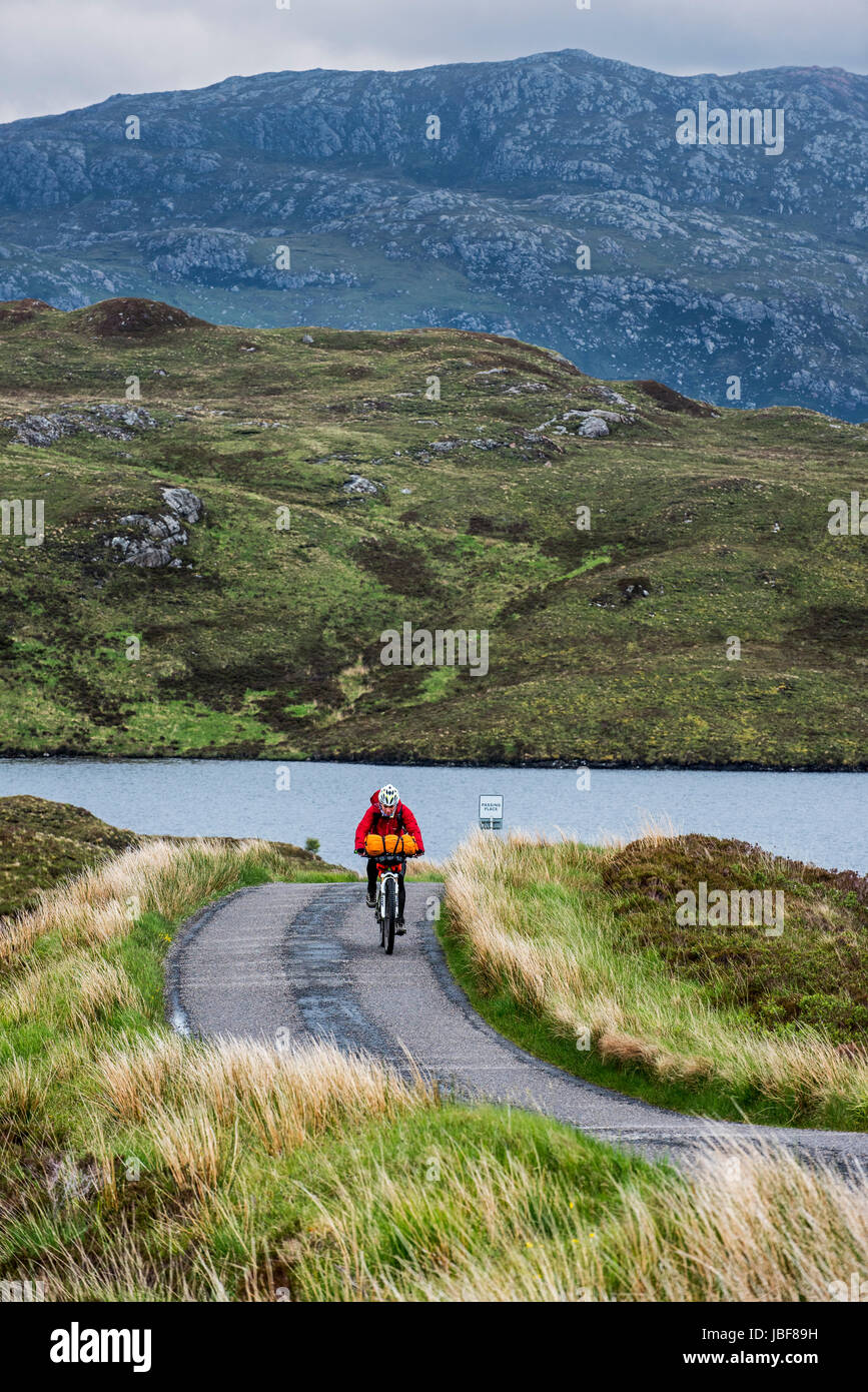 Ciclista solitario ciclismo por las Tierras Altas Escocesas en bicicleta de touring cargados a lo largo de vía única carretera en mal tiempo lluvioso, Scotland, Reino Unido Foto de stock