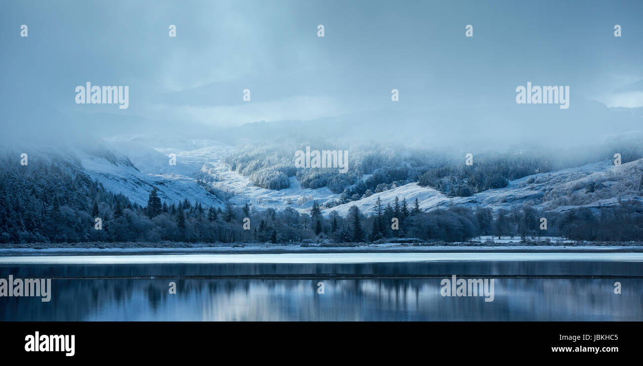 Escena de Invierno en Loch Carron, Torridon, Escocia, con colinas cubiertas de nieve se refleja en el lago en una fría mañana de invierno, Chubascos de nieve en la distancia Foto de stock