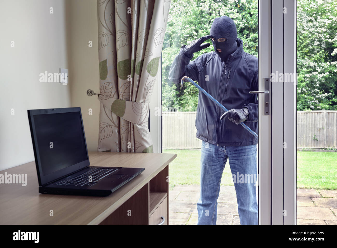 Ladrón entrando en una casa a través de una ventana de puertas de patio con una palanqueta para robar un ordenador portátil desde un escritorio de oficina Foto de stock
