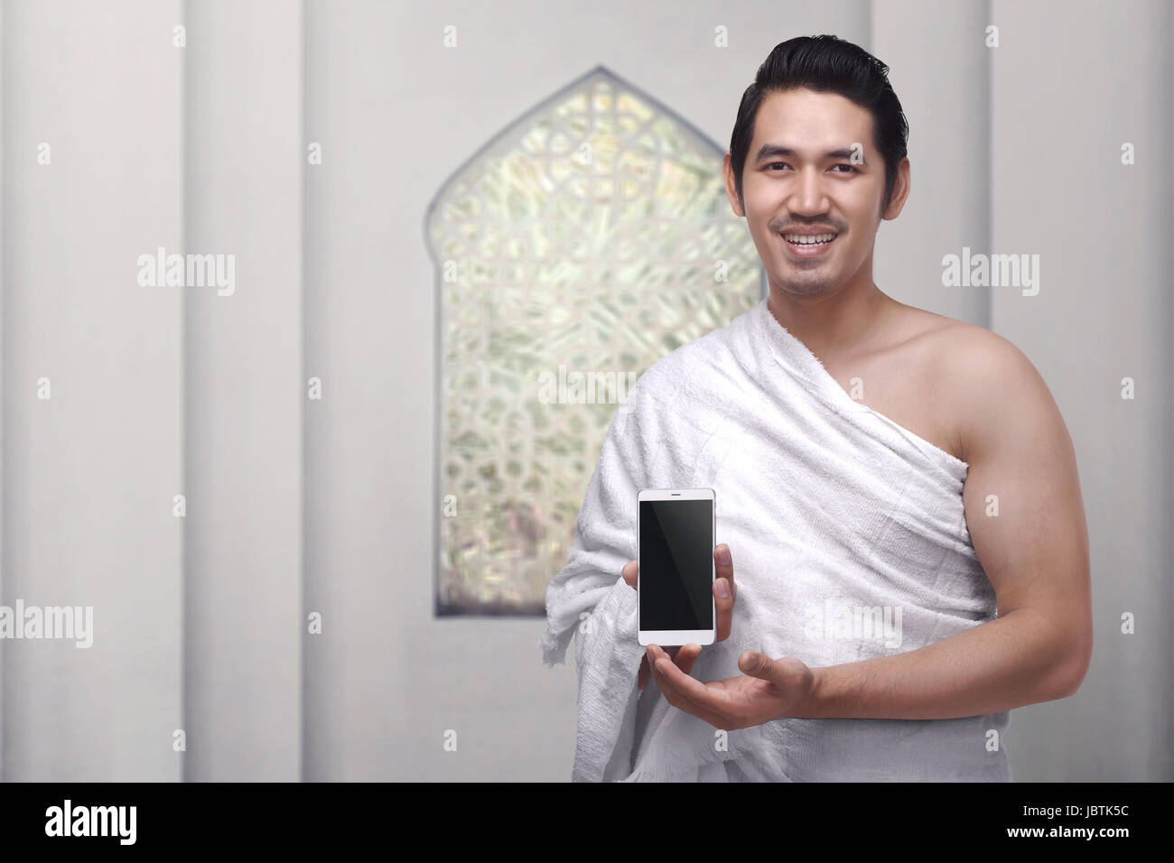 Guapo asiático musulmán vistiendo ropa de ihram holding móvil contra la pared blanca de fondo Foto de stock