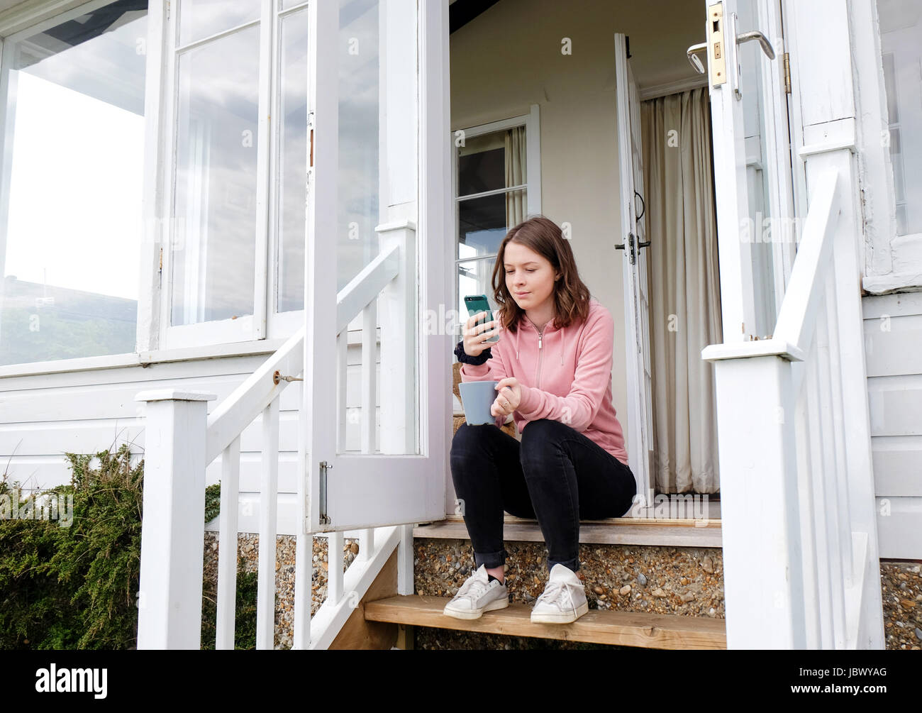 Mujer joven sentada en el porche escaleras con taza de café mirando el smartphone Foto de stock
