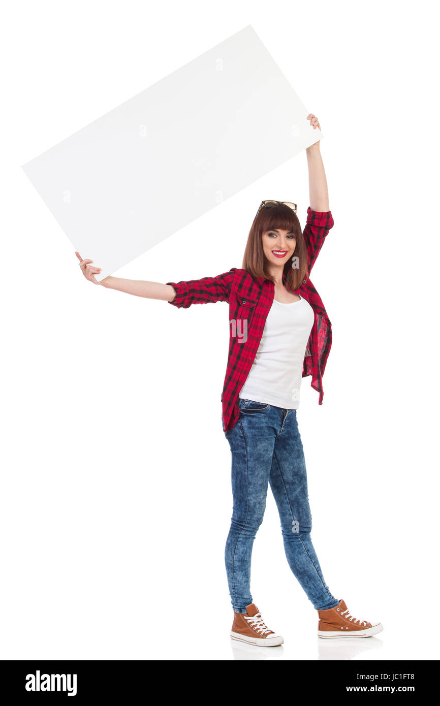 Mujer joven en rojo camiseta de leñadores, vaqueros y zapatillas marrón está de pie, sujetando la etiqueta en blanco blanco por encima de su cabeza y mirando a la cámara. Leng completo Foto de stock