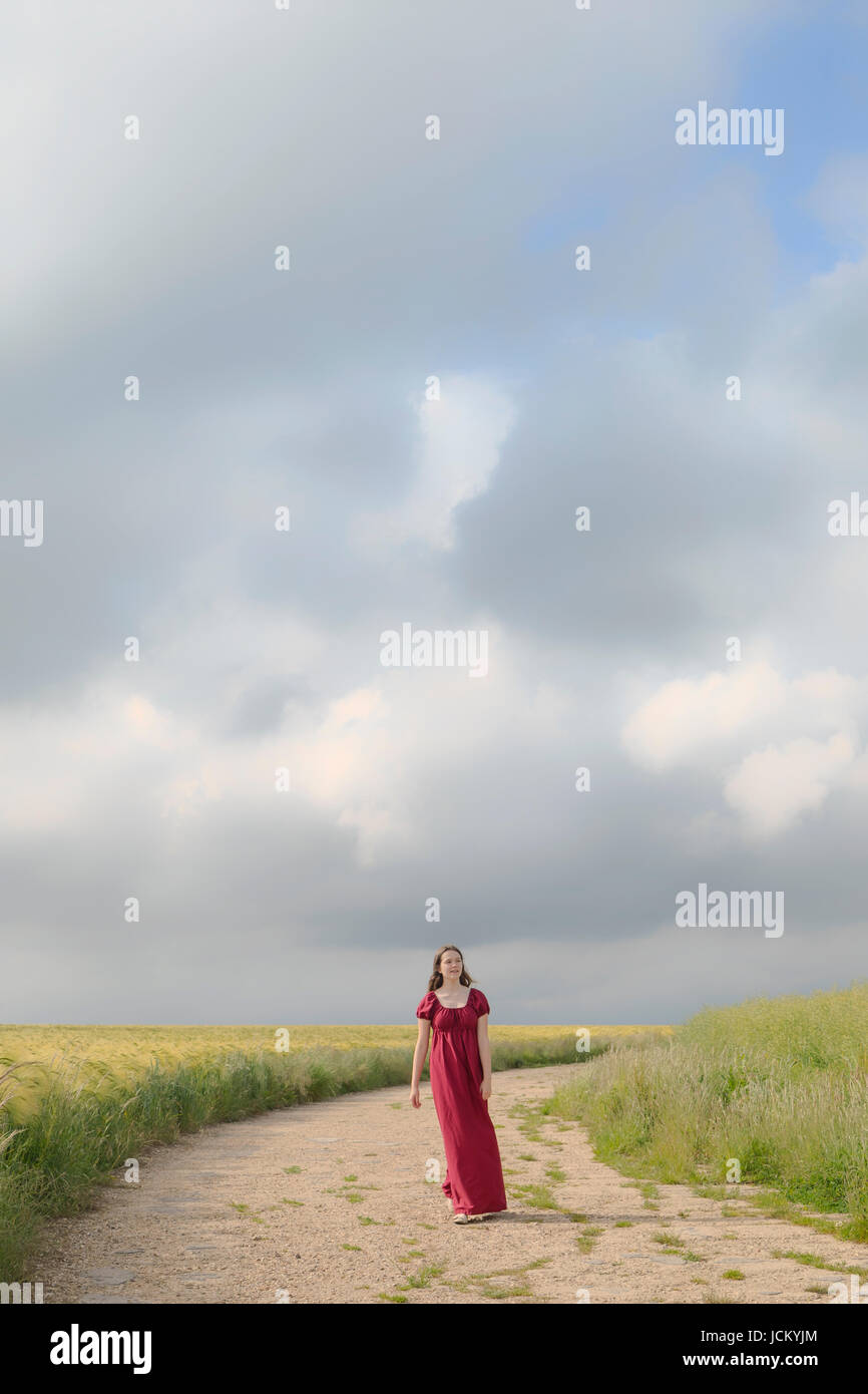 Una mujer en un vestido rojo está caminando por un sendero a través de sembrados Foto de stock