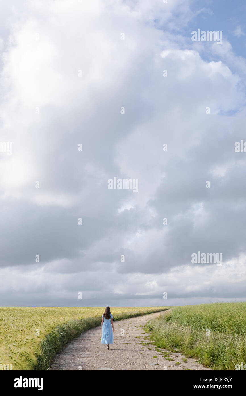 Una mujer en un vestido azul está caminando por un sendero a través de sembrados Foto de stock
