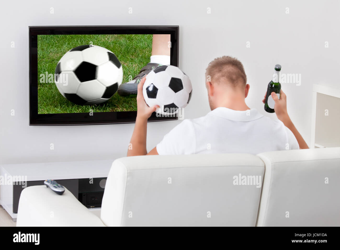 Emocionados hinchas de fútbol viendo un por televisión sosteniendo un balón de fútbol por de su como él está sentado en un cómodo sofá en su sala de estar
