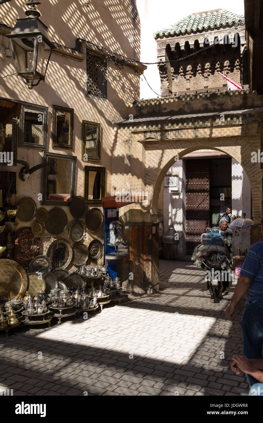 Latón y bandejas de metal, teteras y otros bienes de fantasía en la calle fuera de una pequeña tienda en Fez, Marruecos. Un hombre guía un asno cargado anteriormente. Foto de stock