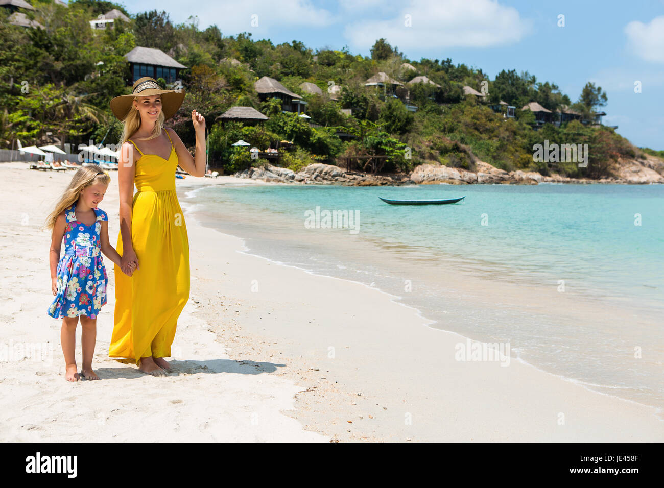 Hermosa mujer rubia en Vestido túnica amarilla larga con su bonita pequeña  hija en azul vestido floral al caminar por la playa. Ellos tomados de la  mano y hablar Fotografía de stock -