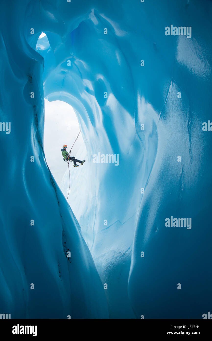 Un escalador de hielo en una chaqueta verde y naranja casco rappeles pasado una entrada grande redondeada a una cueva de hielo en el Glaciar Matanuska en Alaska. Foto de stock