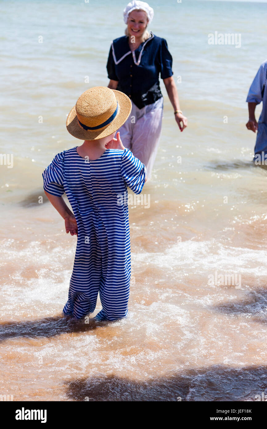 Niños caucásicos, muchacho rubio, de 4-5 años, de pie en el surf en la playa, vestida de azul y sombrero de paja y traje de playa Victoriano blanco. Foto de stock