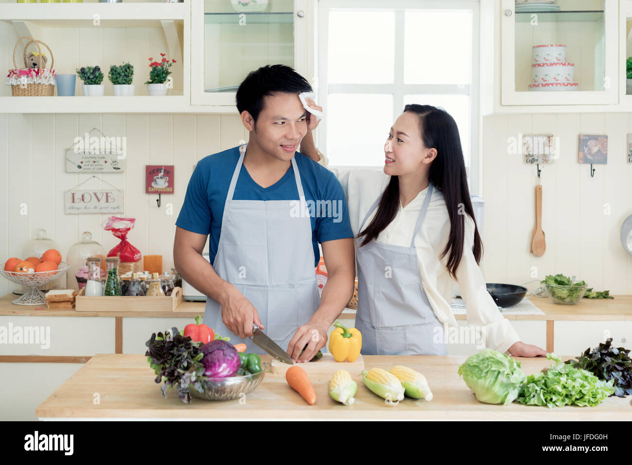 Joven y bella mujer asiática ayudando marido sudor seco su rostro en la cocina de casa. Feliz pareja amor concepto. Foto de stock