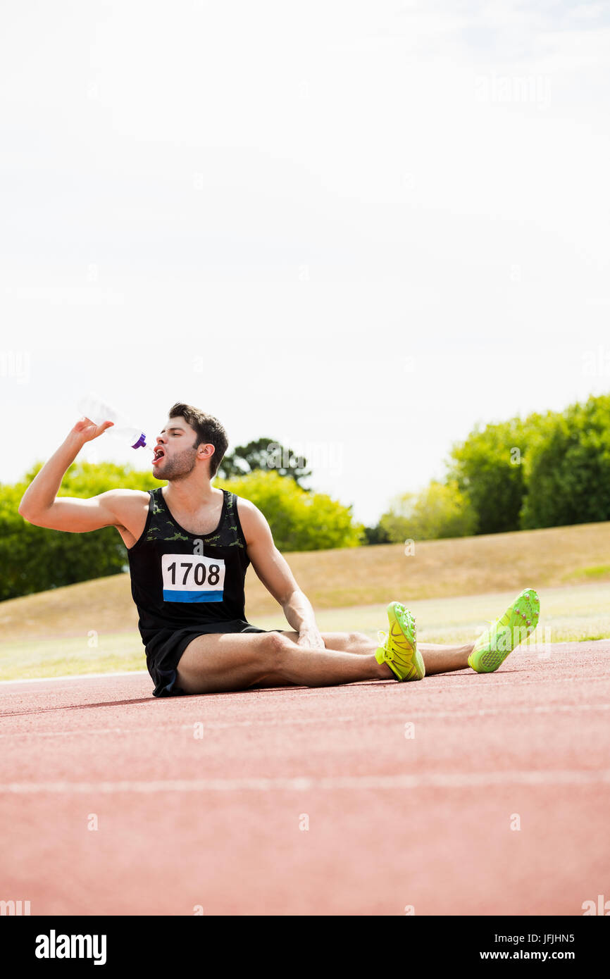 Cansado atleta sentado en la pista de atletismo y agua potable Foto de stock