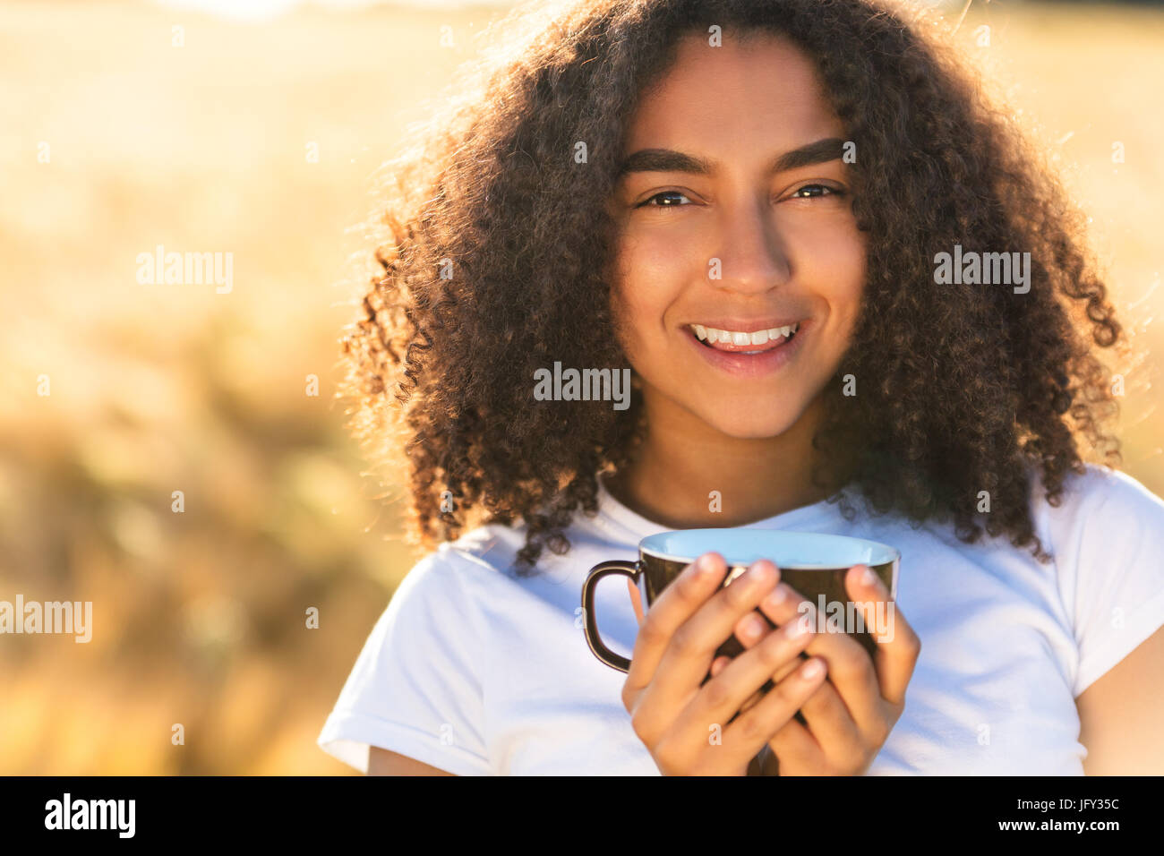 Feliz hermosa chica americana africana de raza mixta femenina adolescente joven sonriendo con dientes perfectos blanca,beber café o té en el exterior Foto de stock