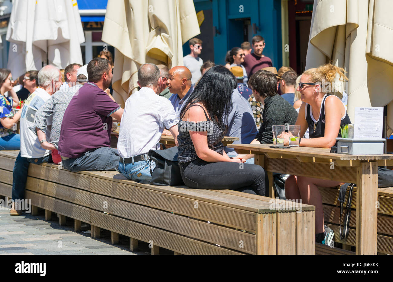 Gente sentada fuera en mesas de madera y bancos en una cafetería y bar en un caluroso día de verano. Foto de stock