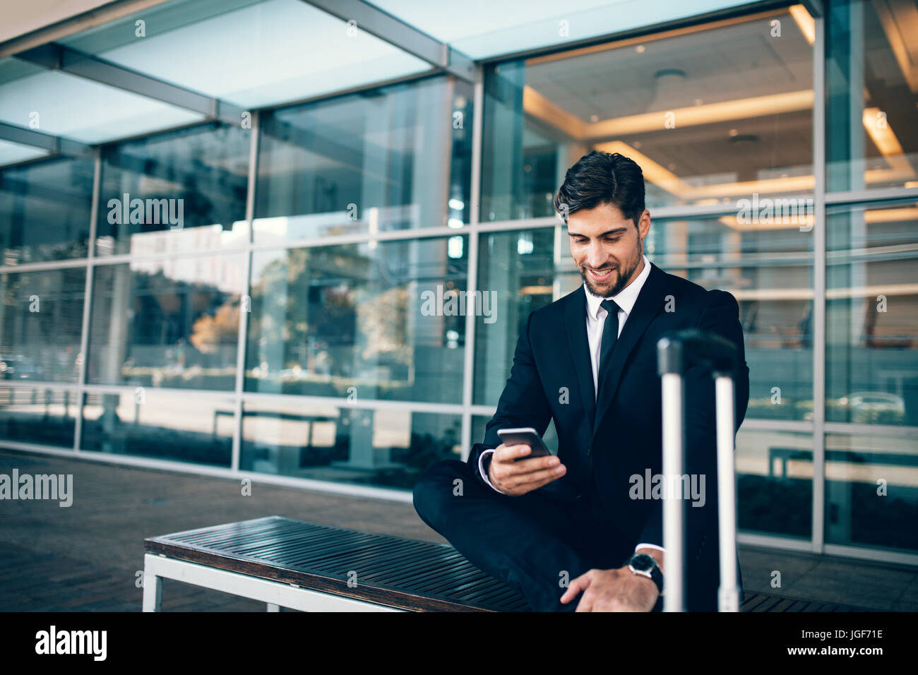 Empresario joven sentado en un banco con la maleta y el uso de teléfonos inteligentes. Empresario del Cáucaso a la terminal del aeropuerto esperando la celebración de un teléfono móvil. Foto de stock