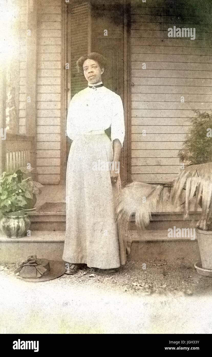 Retrato de cuerpo entero de la afroamericana de pie en frente de un porche, vistiendo un vestido blanco, con su sombrero en el suelo, las plantas en macetas por su lado, con una expresión facial grave, 1920. Nota: la imagen ha sido coloreada digitalmente mediante un proceso moderno. Los colores pueden no ser exactos del período. Foto de stock