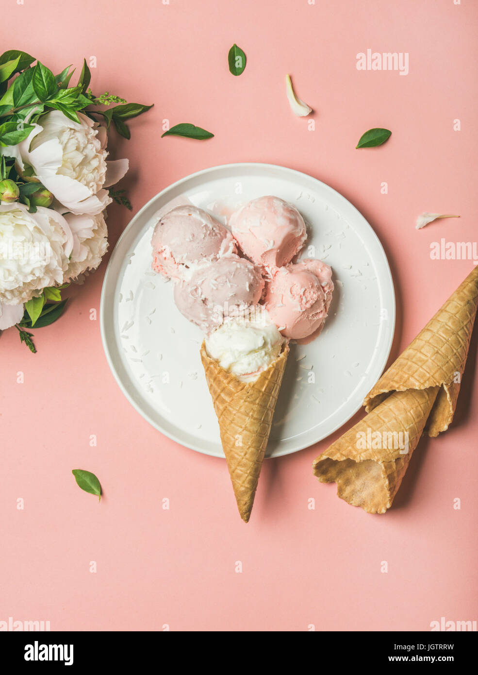 Flatlay de color rosa pastel helado de fresa y coco bocas, dulce conos de placa blanca y peonías blancas sobre fondo de color rosa pastel, vista superior, ve Foto de stock