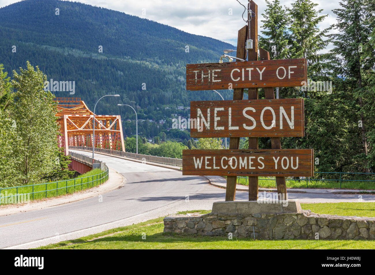 Cartel de bienvenida a la entrada de la ciudad de Nelson, BC, Canadá - con el brazo oeste del puente (Bridge) naranja grande en el fondo Foto de stock