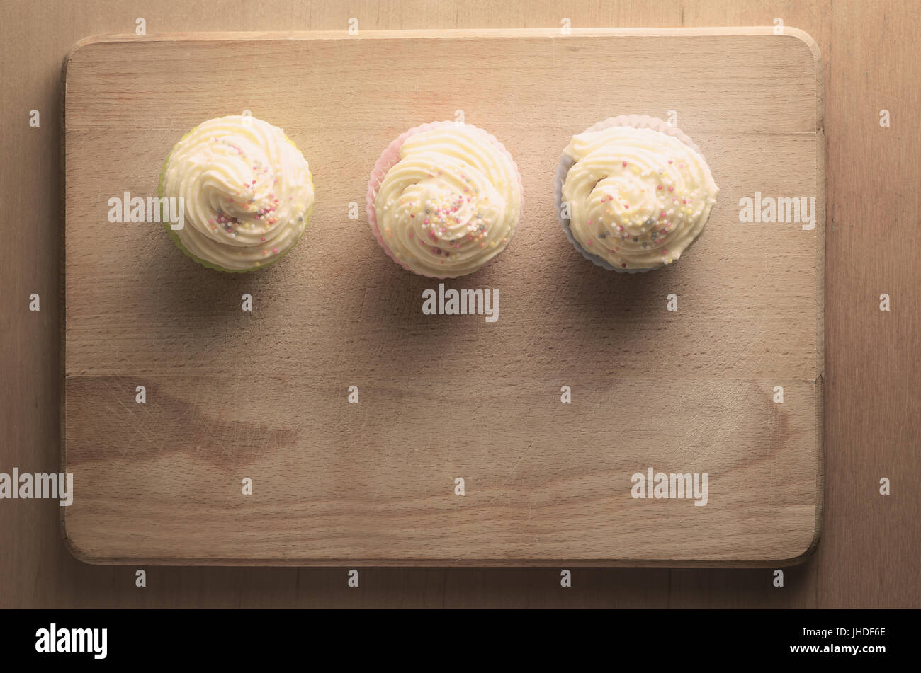 Fotografía cenital de tres pastelitos en una fila en una tabla de cortar de madera, rematada con remolinos de buttercream guinda y coloridos sprinkles. Tonos retro. Foto de stock
