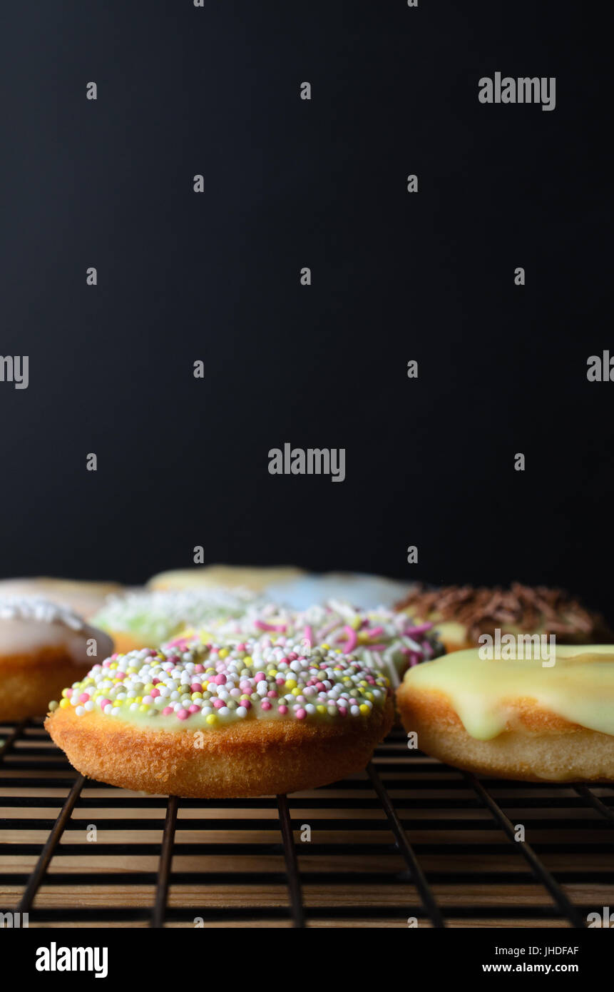 Mini donut pasteles decorados con una gran variedad de sprinkles, sobre una rejilla metálica negra con mesa de madera debajo. Antecedentes de la pizarra negra provi Foto de stock