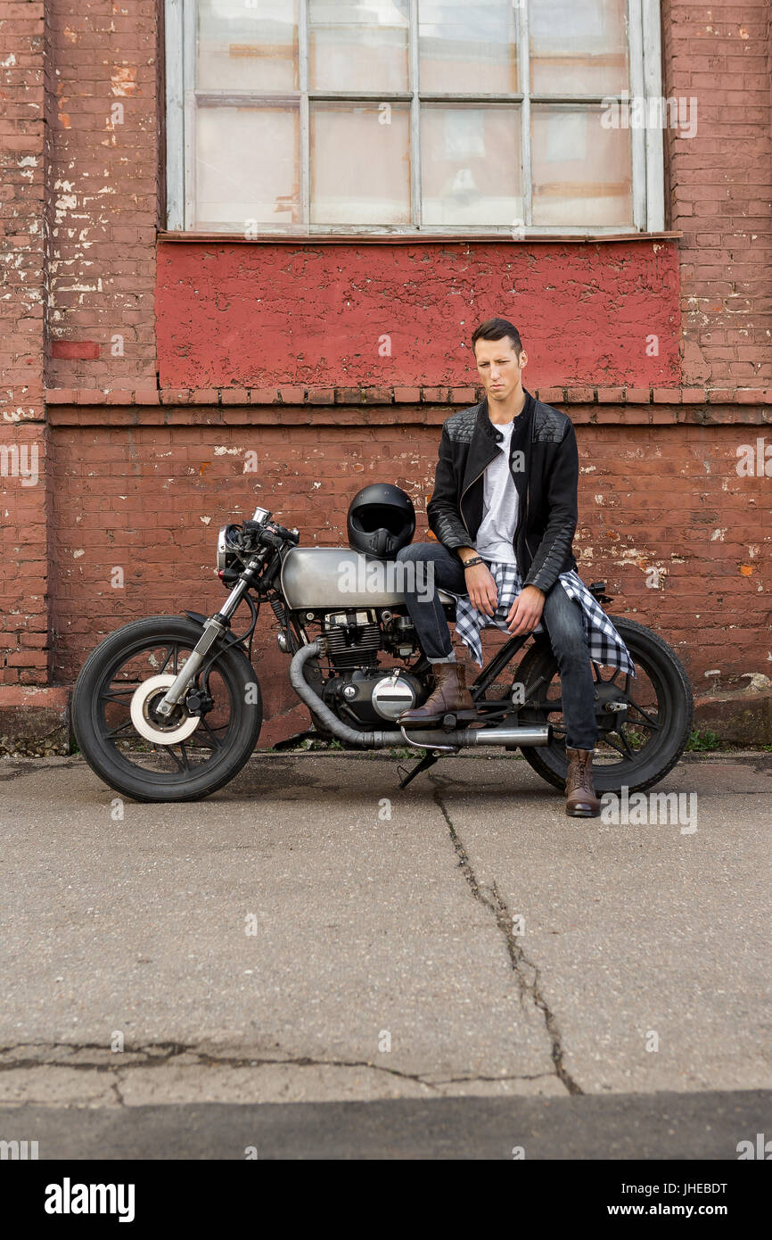 Guapo rider biker chico de la chaqueta de cuero negro, pantalones vaqueros  y las botas sentarse en estilo clásico cafe racer moto. Moto custom made en  vintage garaje. Bruta Fotografía de stock -