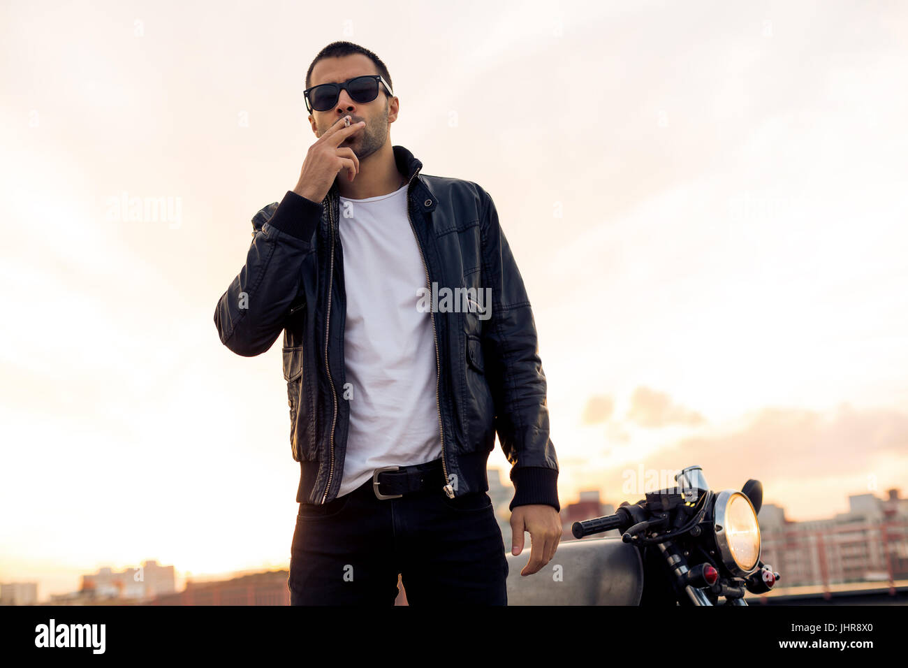 Hermosa feliz Guy Rider con la barba y el bigote en negro biker chaqueta,  jeans y gafas de sol de moda fumar cigaret cerca de estilo clásico café  racer mo Fotografía de