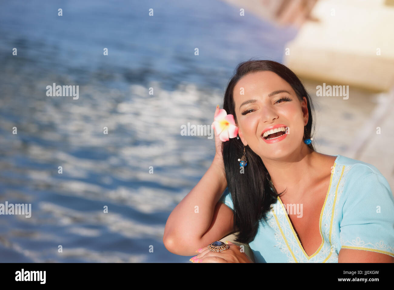 Mujer alegre y al aire libre. Feliz hermosa mujer vistiendo bronceado hawaiano flor en el pelo y disfrutar del sol del verano en Seaside Cafe mirando a la cámara Foto de stock