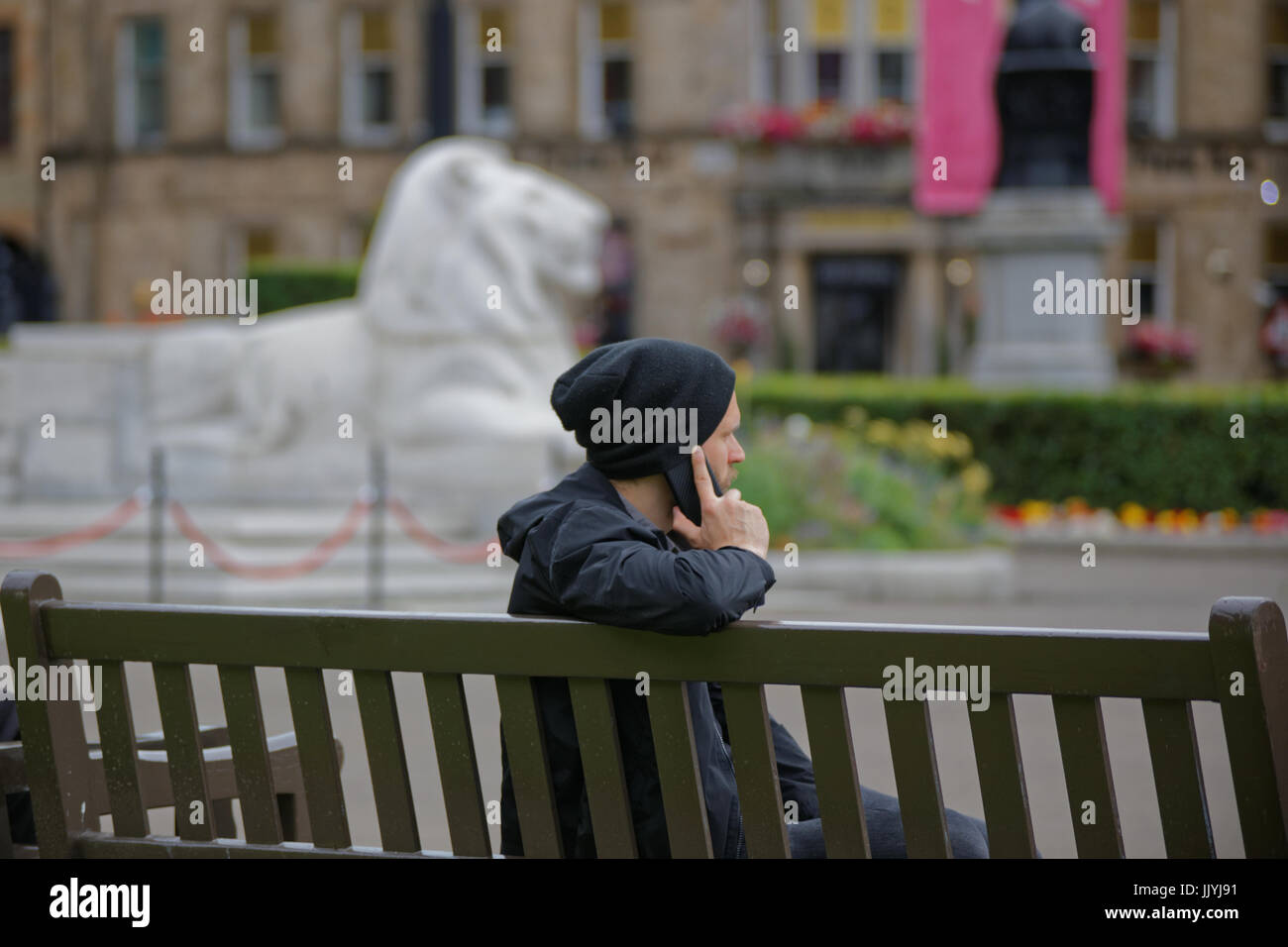 Joven estudiante varón adolescente en el teléfono reflejado en la estatua de león en el fondo detrás de lado o sentado sobre la banqueta om Glasgow George Square Foto de stock