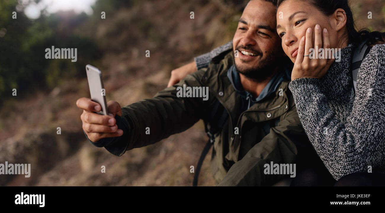 Disparo horizontal de amorosa pareja joven en alza teniendo un autorretrato con el smartphone en el exterior. Excursionista relajada pareja de vacaciones teniendo selfie con cel Foto de stock