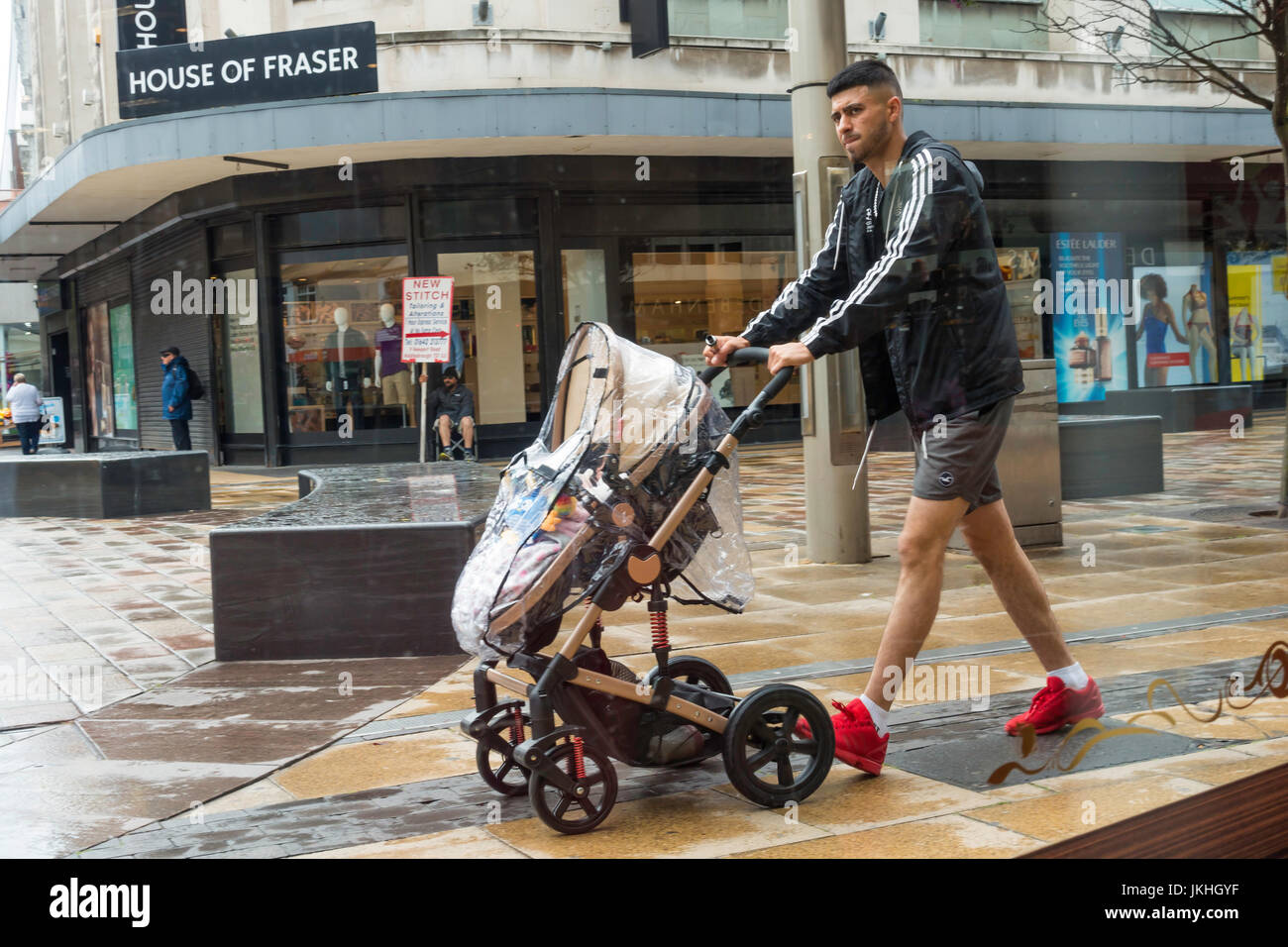 Un joven hombre vestido con pantalones cortos y zapatos rojos empujando un  cochecito de bebé en la lluvia en el centro de la ciudad de Middlesbrough  de Inglaterra Fotografía de stock -
