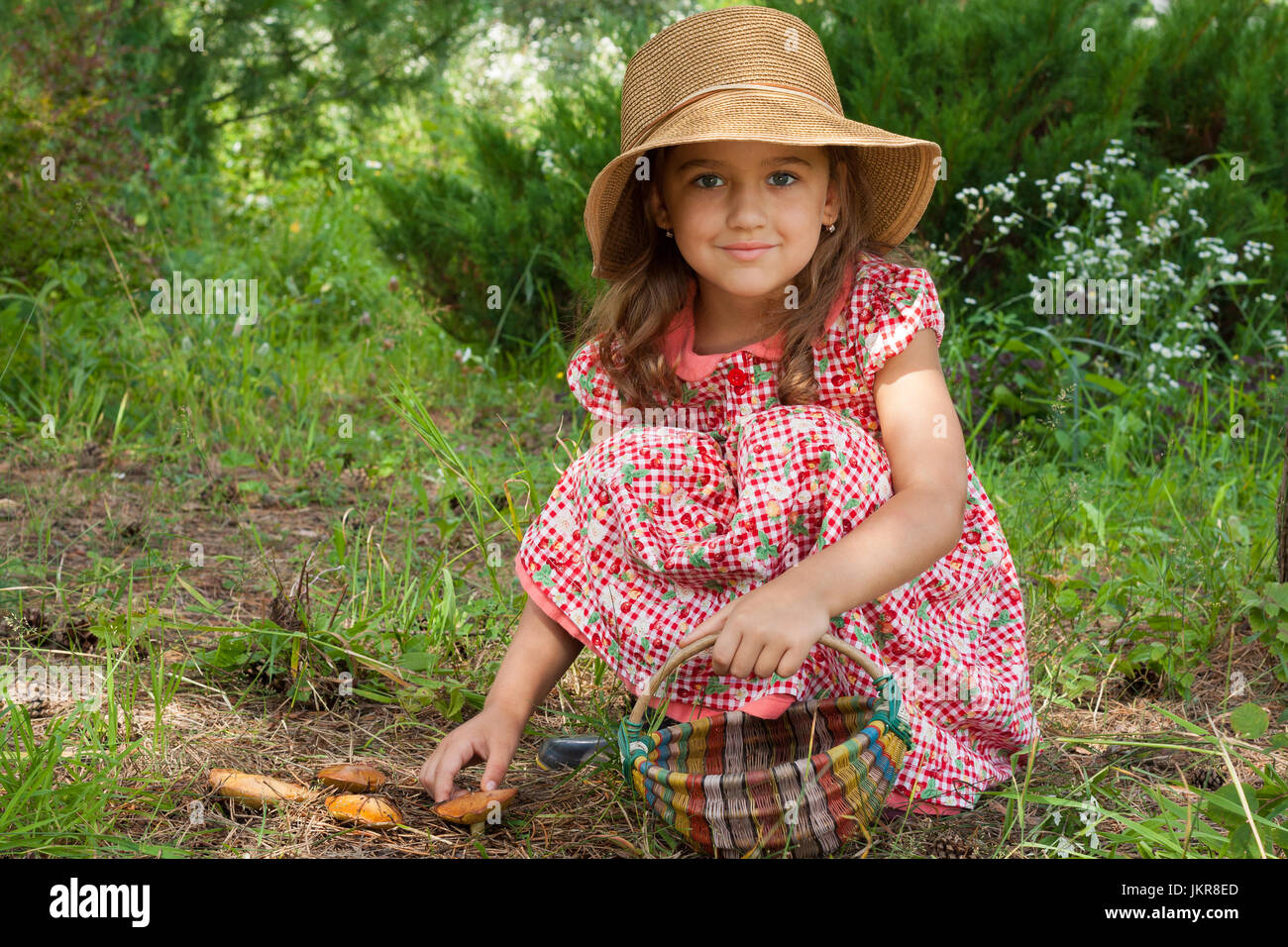 Adorable pequeño de seis años de edad, niña rusa en canasta de mimbre con sombrero en mano encantados de setas suillus, Semi-sentada y mirando a la cámara W Foto de stock