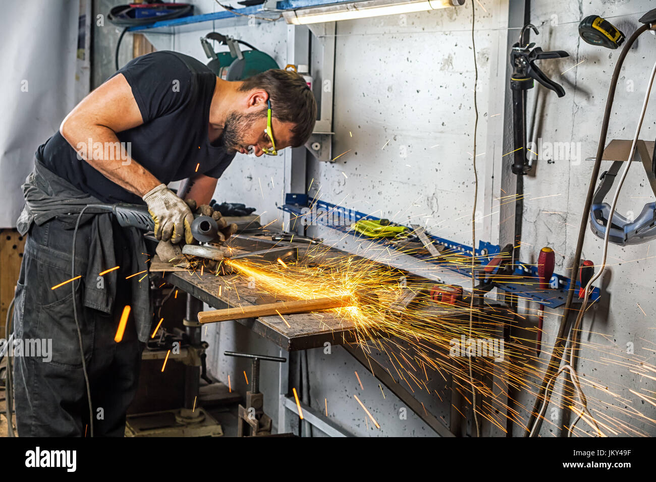 Un joven soldador en una camiseta negra, gafas y guantes de construcción amoladora una angular de metal en el taller,alrededor de montón herramientas para wo Fotografía de stock -