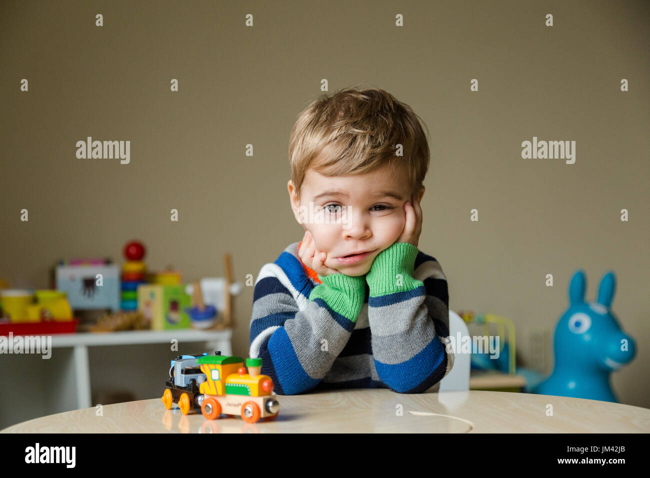 Un niño niño sentado en su sala de juegos se apoya en sus codos sobre una mesa y squishes sus mejillas con una mirada de tristeza en su rostro. Foto de stock