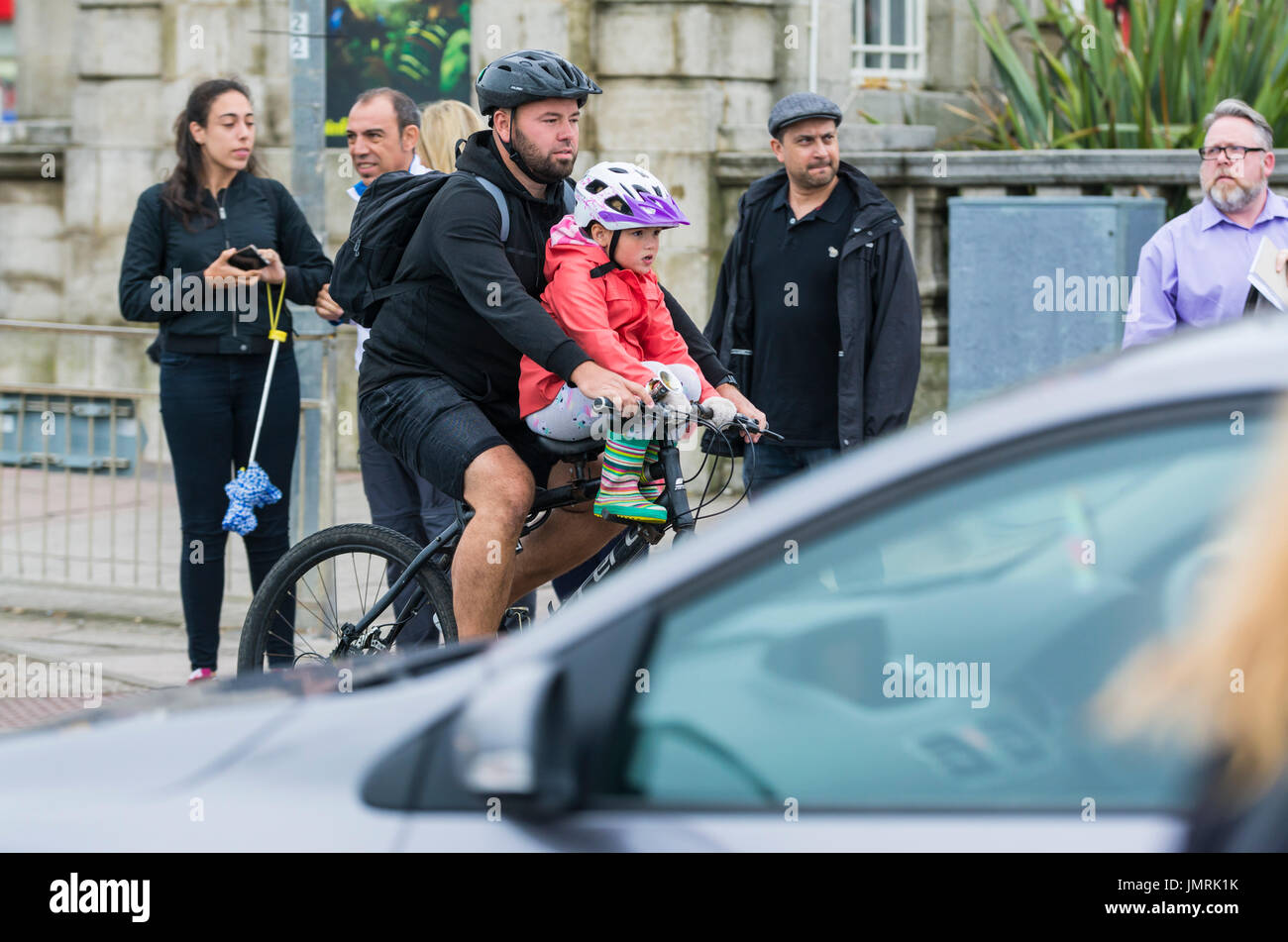 Ciclista con un niño en una carretera muy transitada. Hombre y niño en una bicicleta con cascos. Foto de stock