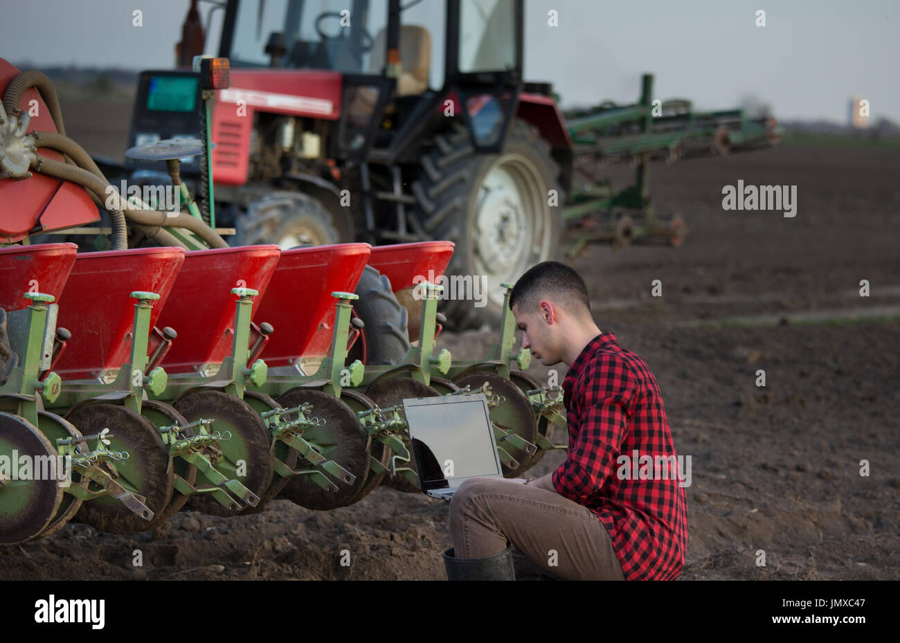 Joven agricultor con laptop en cuclillas en el campo en la parte delantera del tractor con equipo de siembra. Tecnología inteligente en la agricultura Foto de stock