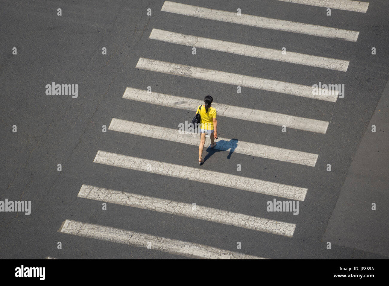 Vista aérea de la mujer en la parte superior amarilla caminando con sombra a través de líneas de cruce peatonal de una amplia calle Foto de stock