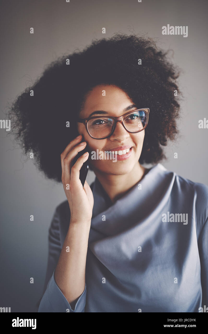 Sonriente joven empresaria africana con gafas y aislada en una oficina hablando por un teléfono móvil Foto de stock