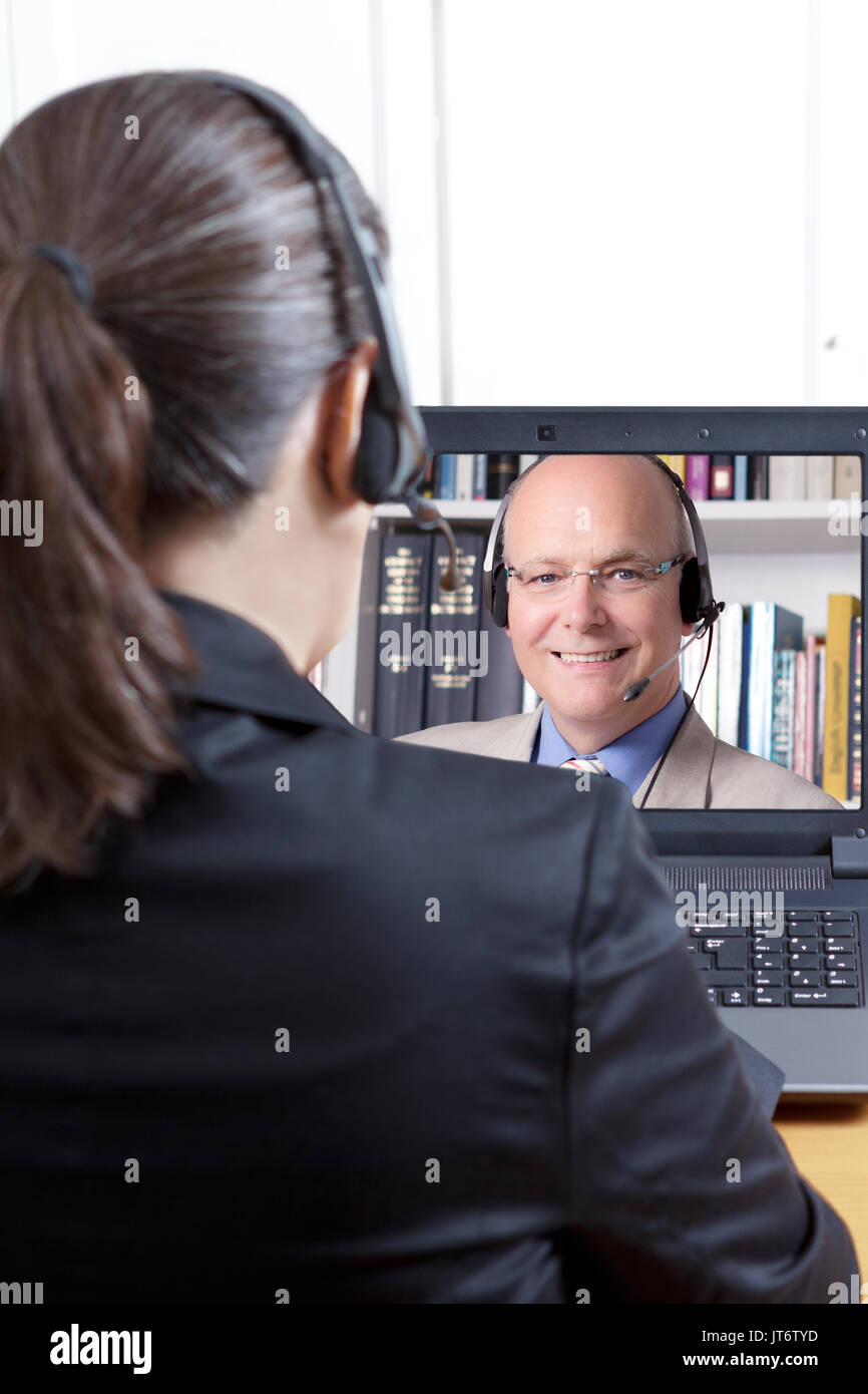Vista trasera de una mujer en un blazer negro con auriculares delante de un ordenador, escuchando a un experimentado profesor de inglés, o e-learning telelearning Foto de stock
