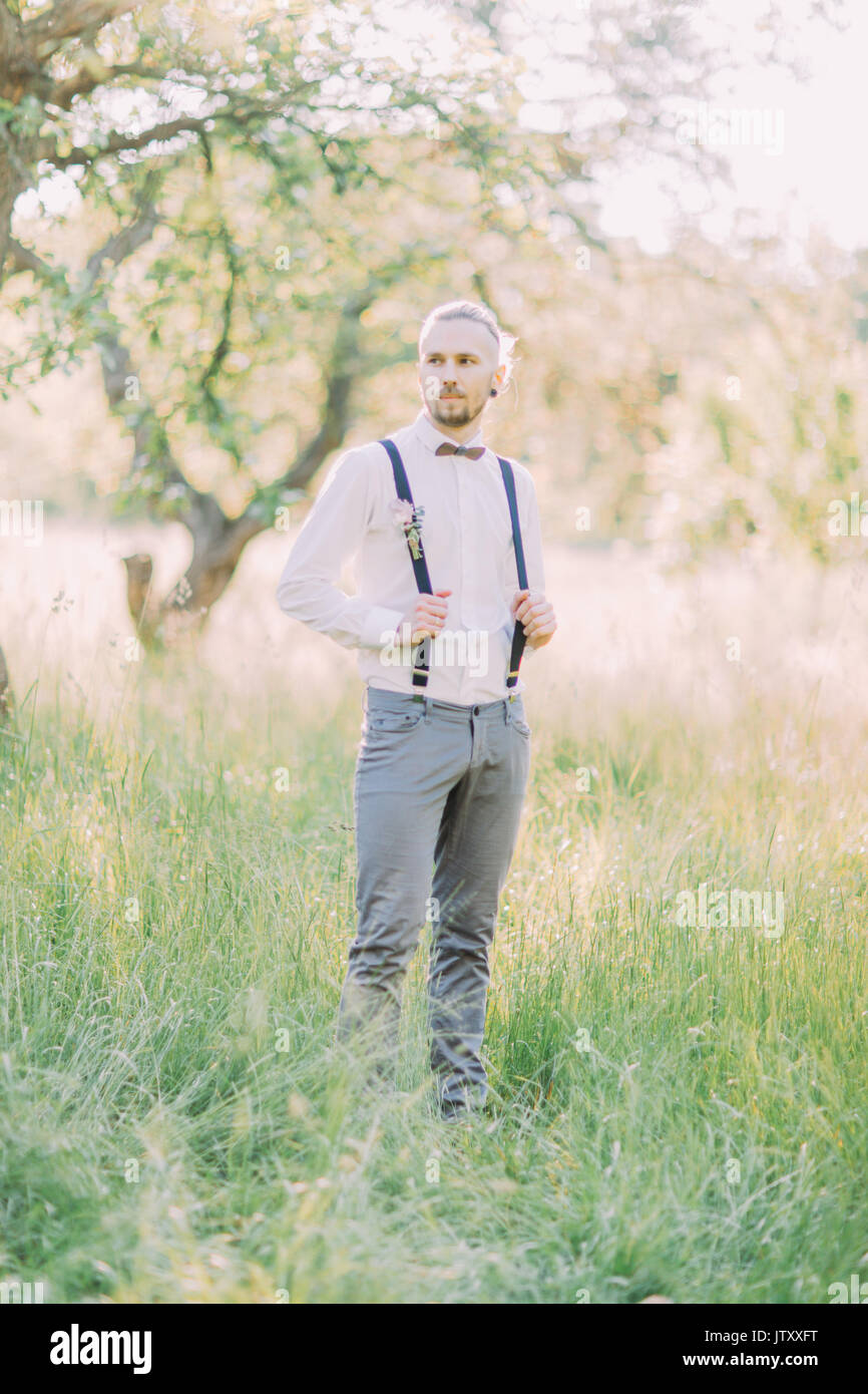 El novio en el pantalón gris y camisa blanca sosteniendo sus tirantes  negros en el verde campo de primavera Fotografía de stock - Alamy