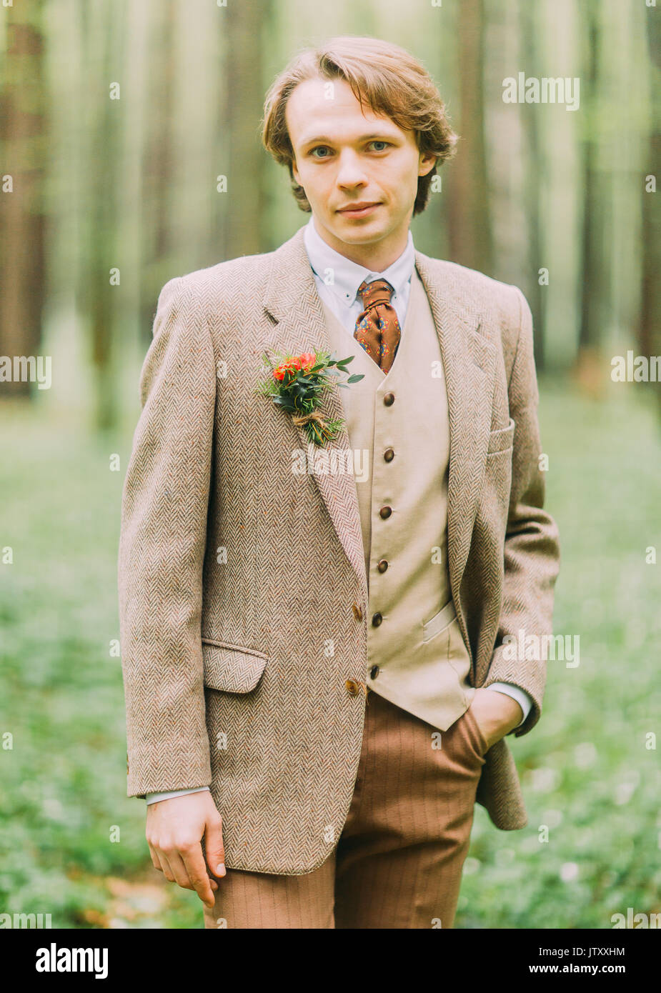 El hombre en el traje vintage, con mini-ramo de flores está anclado en su chaqueta está la cámara y mantener la mano en su bolsillo. Close-up retrato frontal en la madera