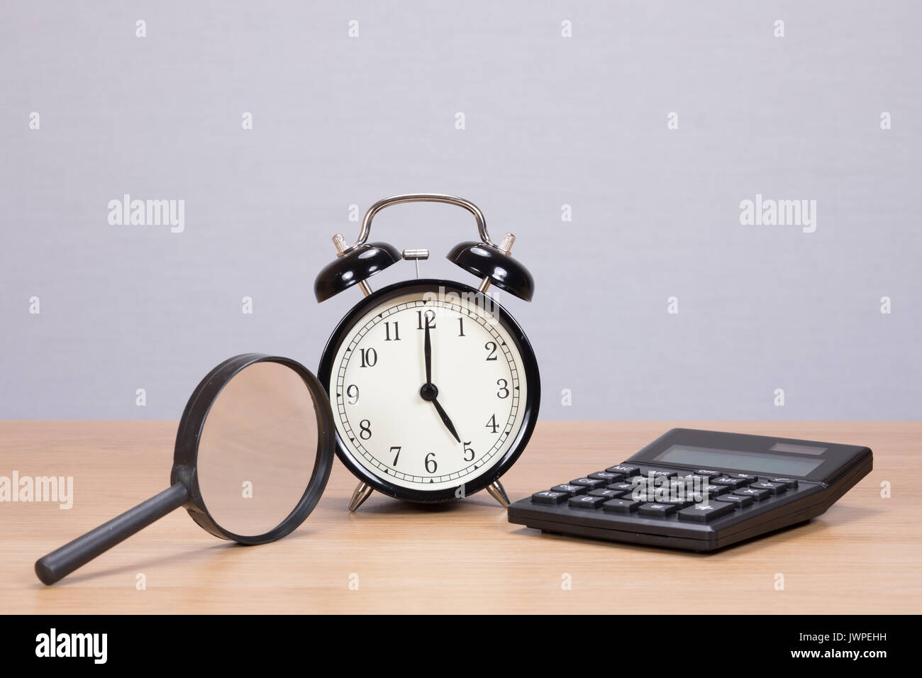 Lupa, calculadora y despertador dispuestos sobre una mesa de madera con copia espacio en una pared gris detrás Foto de stock