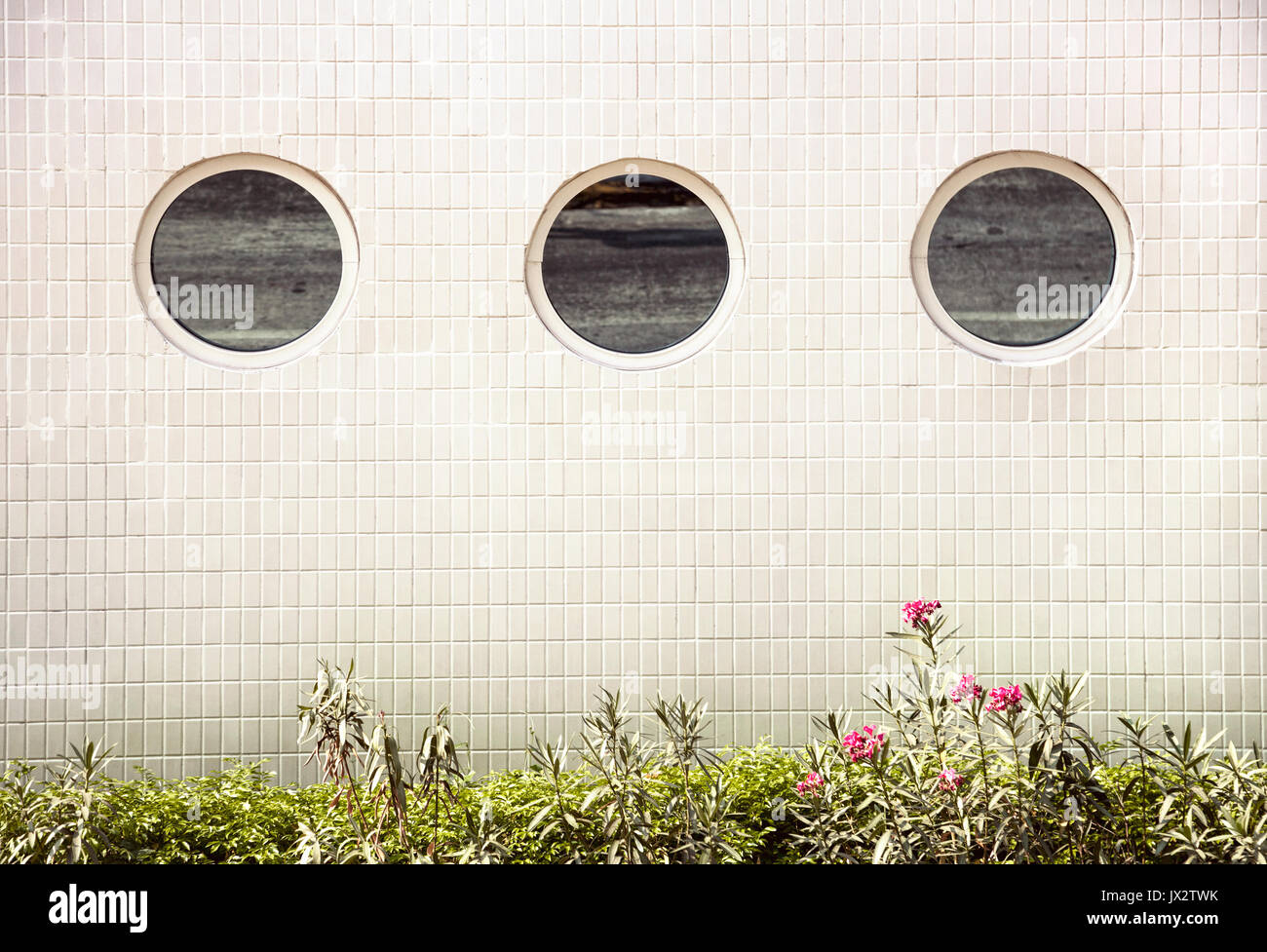 Espejo redondo de 3 ventanas en una fila en la pared del edificio de baldosas blancas y fondo verde hierba y flores debajo Foto de stock