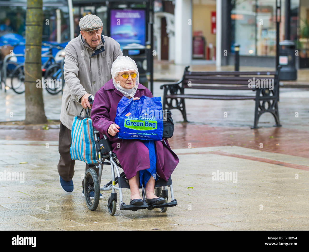 Pareja de ancianos de compras en la lluvia, una empujando a los demás en una silla de ruedas, llevando un Tesco "bolsa de por vida". "La pequeña bolsa verde'. Viejo deshabilitada la lluvia. Foto de stock