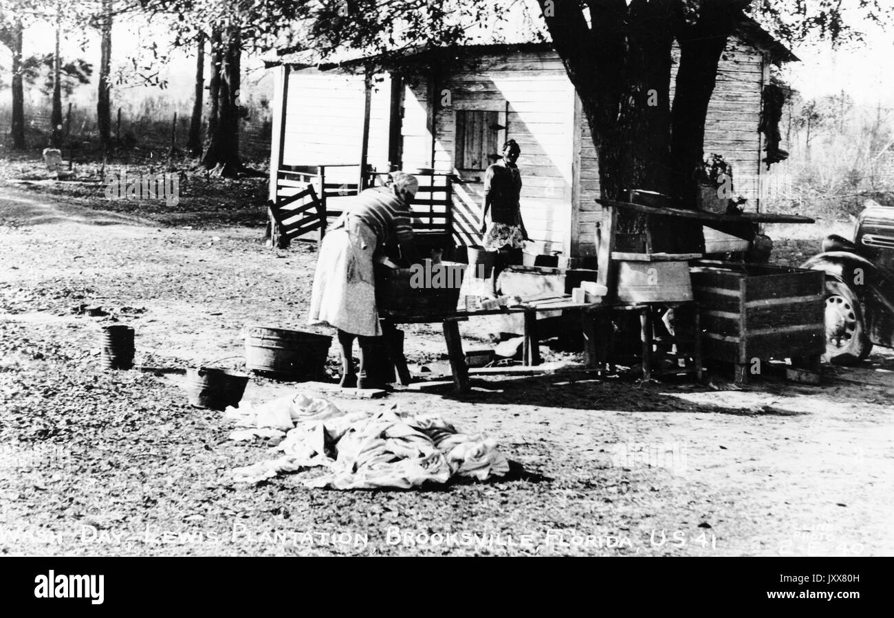 Dos trabajadoras afroamericanas, una llevando una cubeta, una lavando un objeto en un cubo grande en un banco, se encuentran en una zona boscosa despejada fuera de una chabolas, en la plantación Lewis en Brooksville, Florida, 1940. Foto de stock