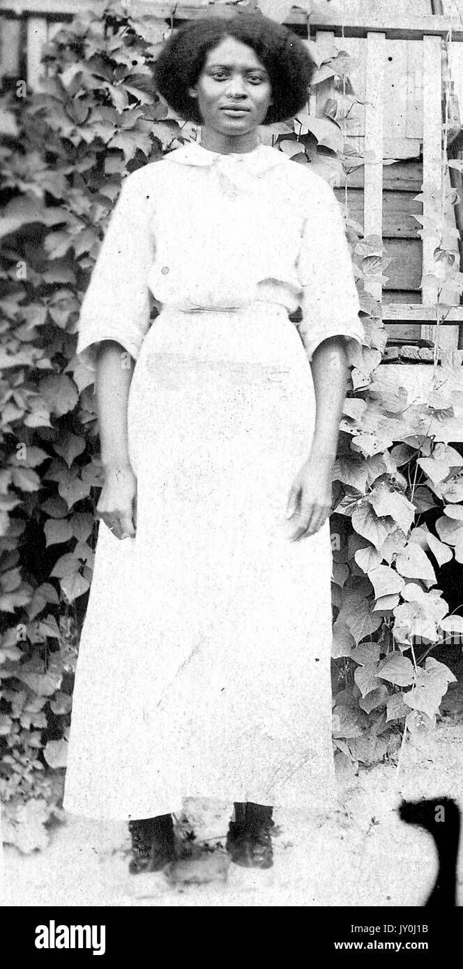 Retrato de una mujer afroamericana de pie frente a un porche cubierto de hiedra, está usando una blusa de color claro y está metida en una falda larga de color claro, está usando zapatos de color oscuro, 1915. Foto de stock