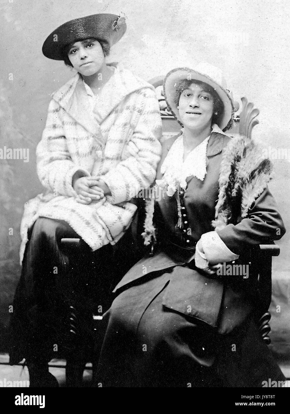 Retrato de dos mujeres afroamericanas sentado en una silla, vistiendo vestidos y sombreros, en un estudio fotográfico, una mujer con una ligera sonrisa, 1915. Foto de stock