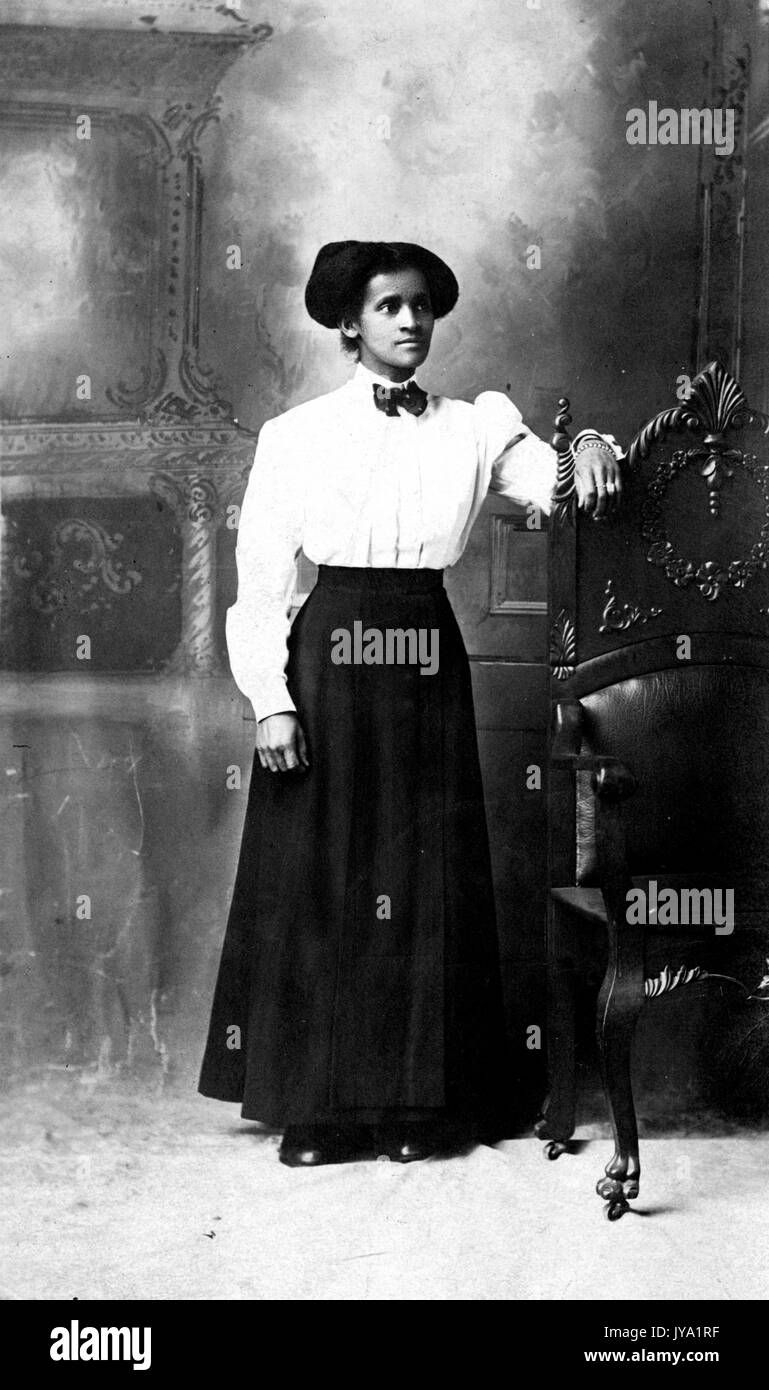 Retrato de una mujer afroamericana de pie delante de un mural decorativo, está descansando una está en una parte posterior de madera alta de una silla, está usando una camisa de color claro con una pajarita y una falda de color oscuro de 1920. Foto de stock
